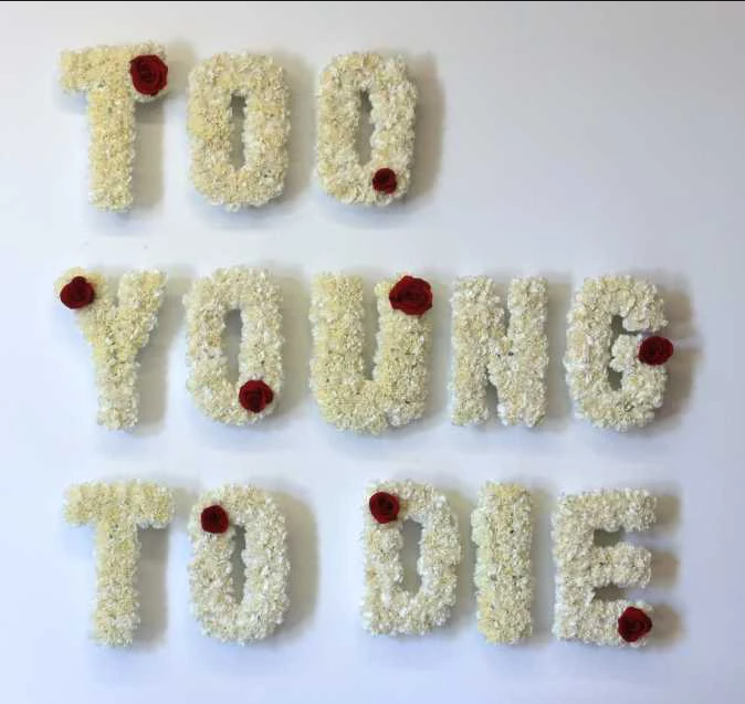 Too Young To Die, 2014. Fresh white carnations, red roses, foam letters. Image courtesy of the artist.