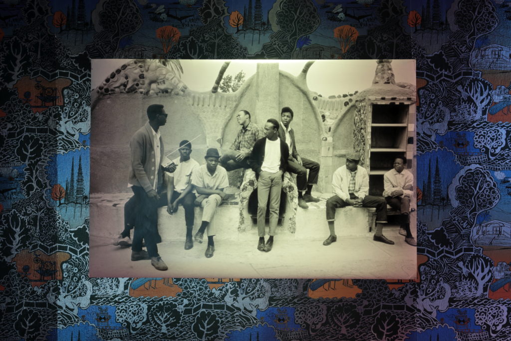 Older photograph of a group of young black men against artist-created wallpaper in A collection of miscellaneous objects on the table in Cauleen Smith's "Give it Or Leave it" Installation