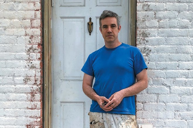 The artist Corin Hewitt stands in front of his studio, which is fronted by a white door and white painted brick. Corin wears a blue shirt.