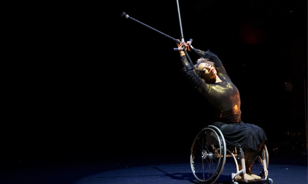 Alice Sheppard, a coffee-skinned multiracial Black woman with short curly hair, reaches her arms overhead. Her silver crutches extend her reach even further, crossing just above her hands. They glint under the stage light, along with her wheelchair and golden long-sleeved costume. Her eyes are closed as she takes in the moment. Photo by Mengwen Cao, during a performance of Where Good Souls Fear.